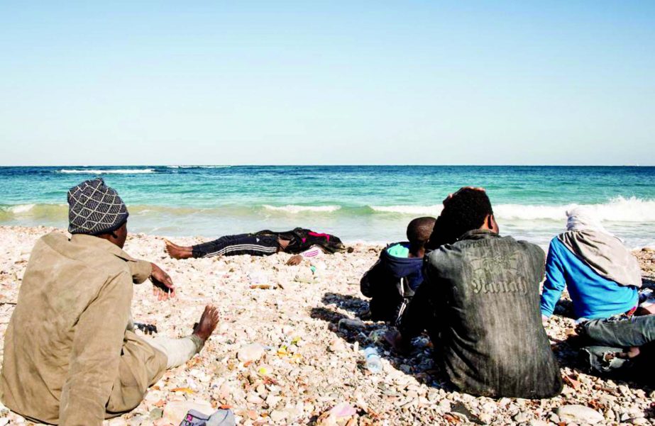The dead body of migrant is seen as other surviving migrants, who were on a boat which capsized on Wednesday, sit on a beach after policemen arrested them in Tripoli, Libya.