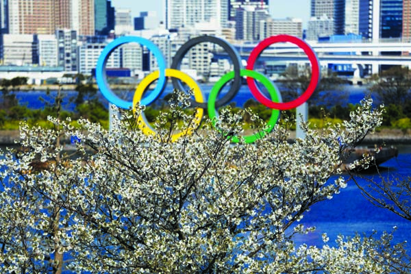 The Olympic rings floating on a barge are seen behind cherry blossoms in the Odaiba section of Tokyo on Wednesday. Not even the Summer Olympics could withstand the force of the coronavirus. After weeks of hedging, the IOC took the unprecedented step of po