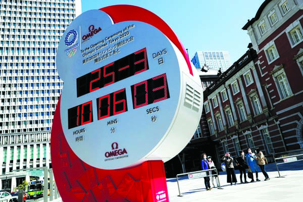 People wearing protective face masks due to the outbreak of coronavirus disease (COVID 19) stand next to an Omega clock, which was previously used as a countdown clock for the Tokyo 2020 Olympic Games and currently displaying current time and date, after