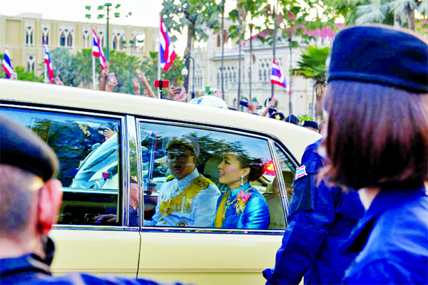 Thailand's Queen Suthida and Prince Dipangkorn Rasmijoti inside a royal motorcade as anti-government protesters (back) hold up the three-finger salute. Two men have been charged over the incident.