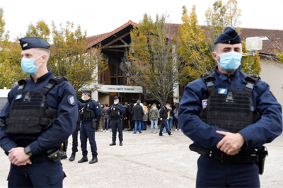 French police officers stand as adults and children gather in front of flowers displayed at the entrance of a middle school in Conflans-Sainte-Honorine.